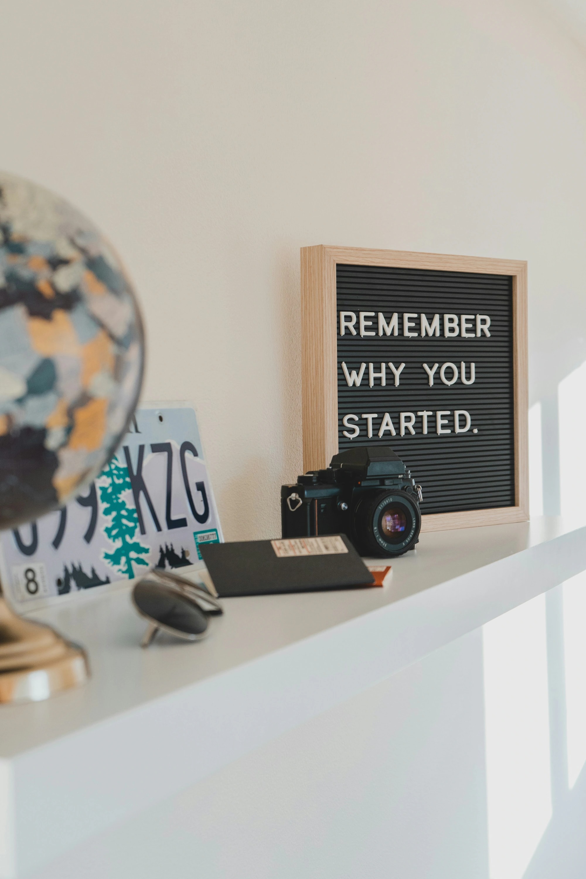 A letterboard reading Remember why you started, beside a camera and globe on a shelf
