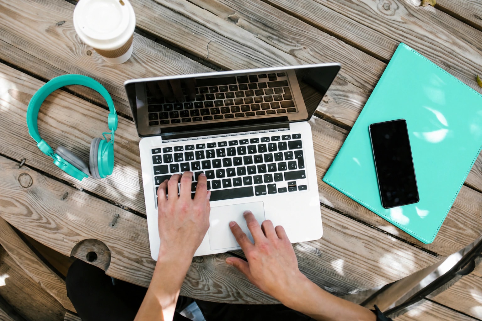 Hands using a laptop on a wooden table beside teal headphones, a mint folder, a phone and coffee