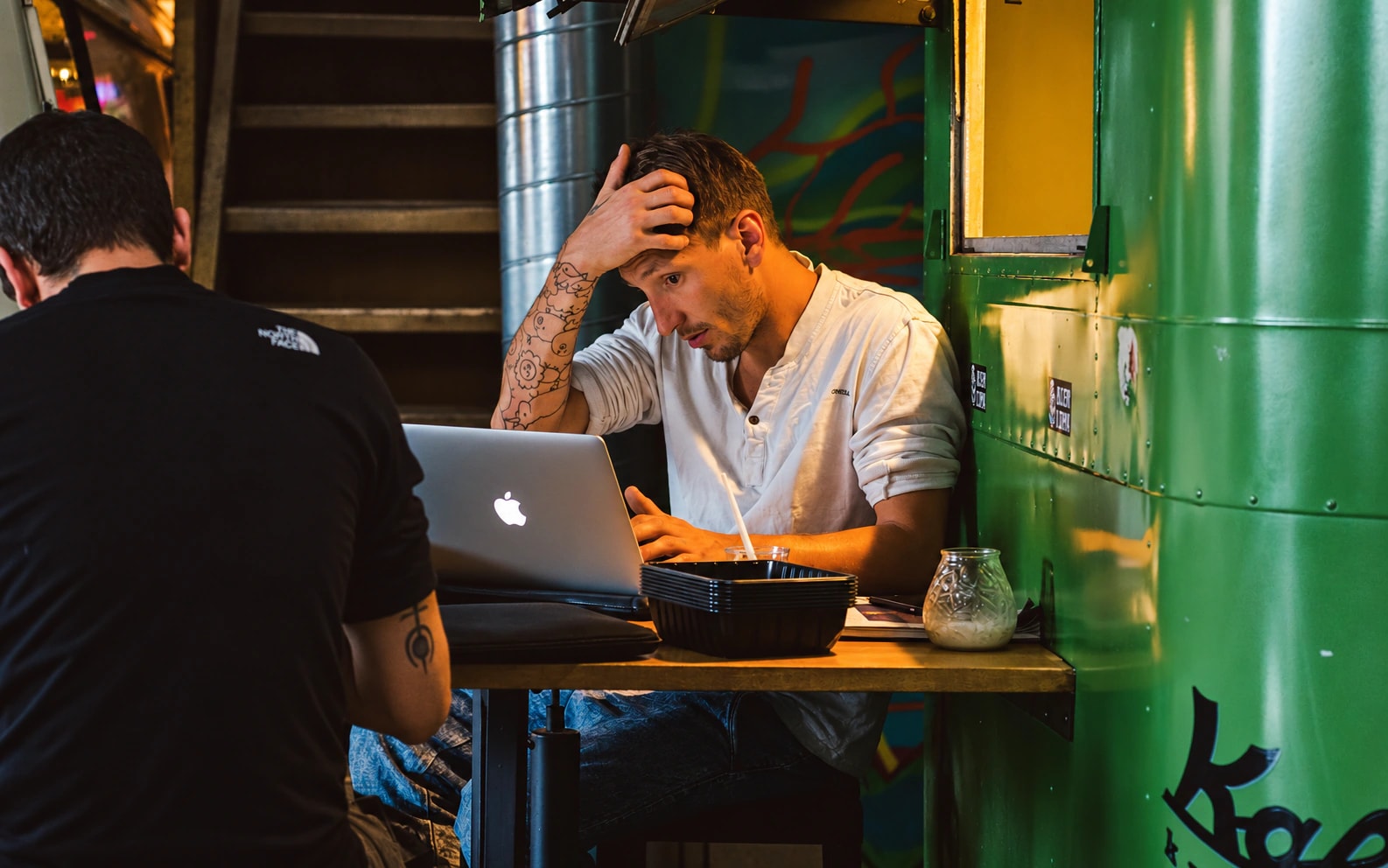 A man using a laptop at a cafe table with one hand resting on his forehead