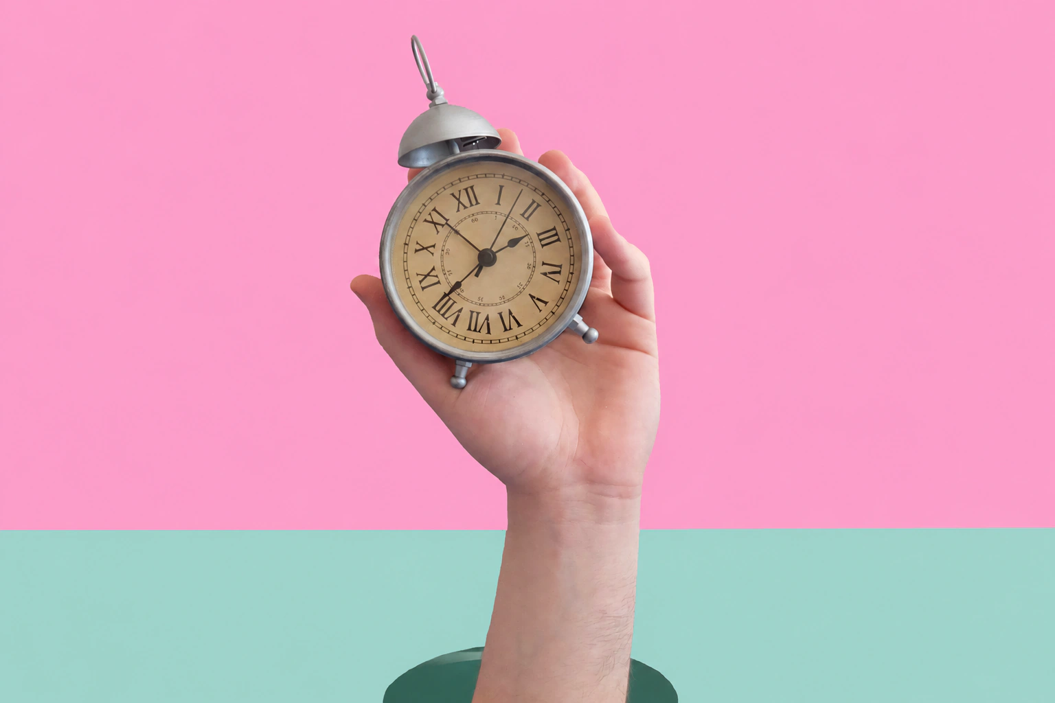 A hand holding a vintage alarm clock against a pink background above a mint green surface