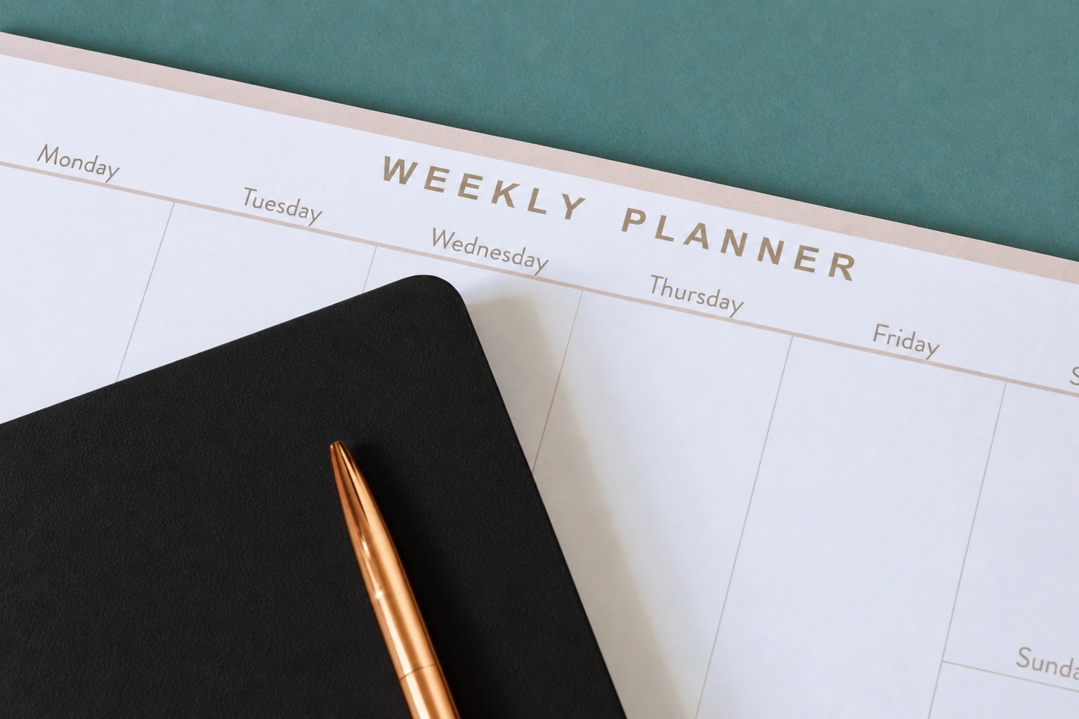 A weekly planner beside a black notebook and copper pen on a muted green surface
