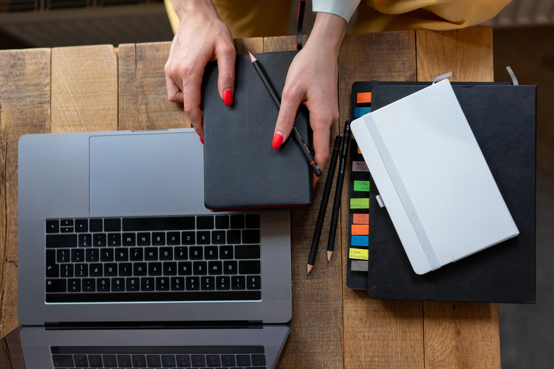 Hands reaching for a notebook beside a laptop and stationery on a desk