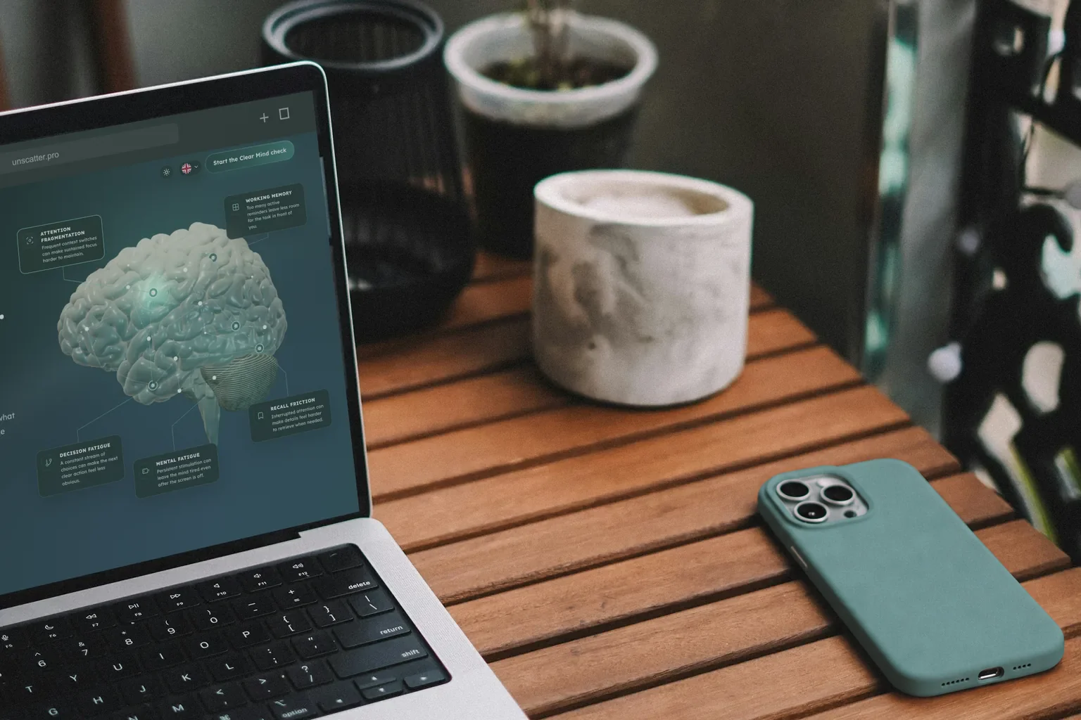 A laptop showing the unscatter homepage on a wooden balcony table beside a plant and phone.